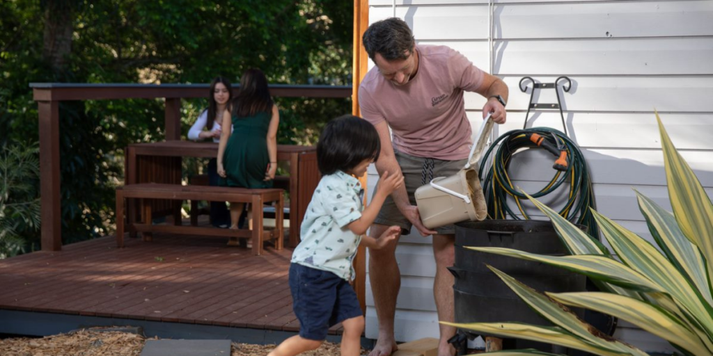 Man emptying compost caddy contents into large worm farm with younger son watching.