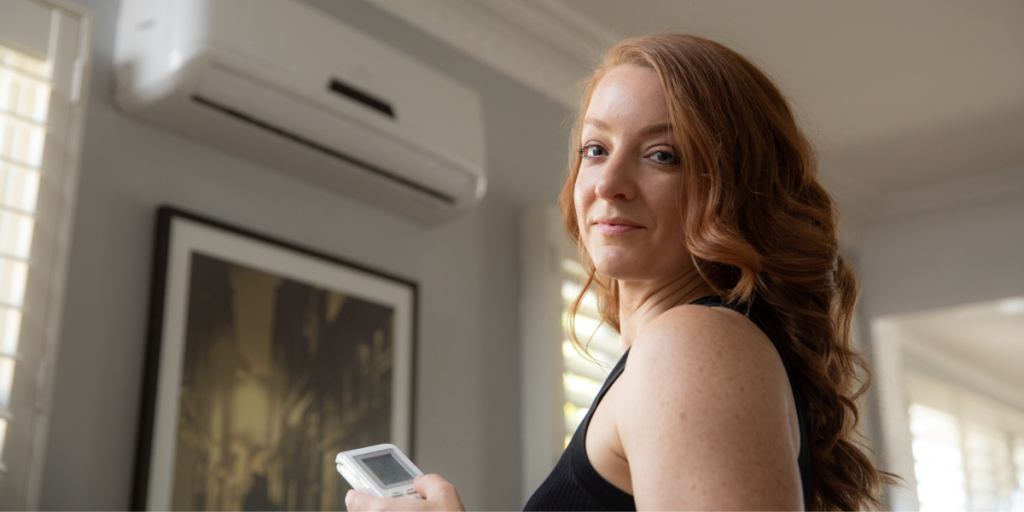 Brisbane resident, Marina Wood, staring into camera smiling as she turns down air conditioning unit with remote.
