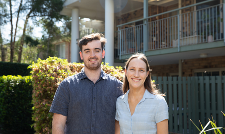 Laura and Finn standing in the yard outside their apartment smiling into the camera.